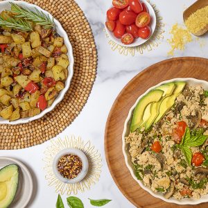 Tofu Scramble with Mushrooms and Basil on a white plate served beside Roasted Potatoes. The background is white marble. Also pictured are diced cherry tomatoes, a half of and avocado, nutritional yeast, red chili pepper and fresh basil leaves.
